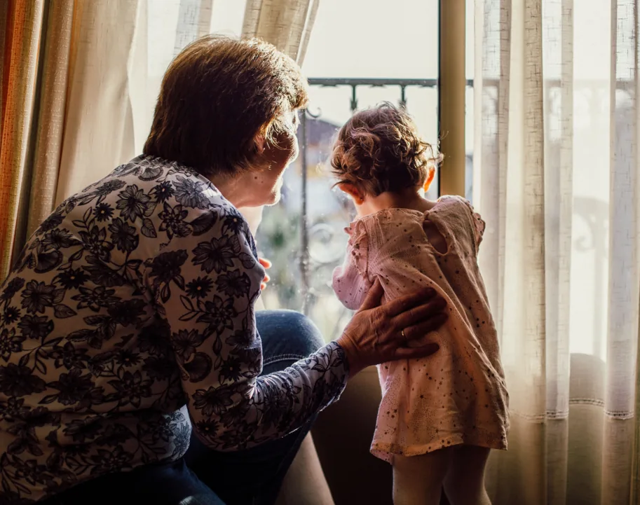 A grandmother and young child looking out a sunlit window together